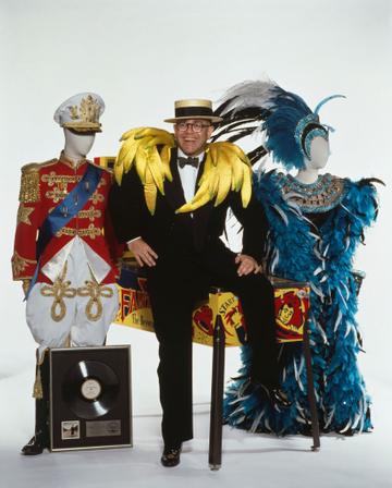 English pop singer Elton John with some of the items he will be putting up for auction at Sotheby's, London, 1988. Among the items are a platinum disc of his album 'A Single Man', a Captain Fantastic and the Brown Dirt Cowboy pinball machine by Bally, and a pair of outlandish stage costumes. John himself is wearing a set of satin bananas over his shoulders. (Photo by Georges De Keerle/Getty Images)